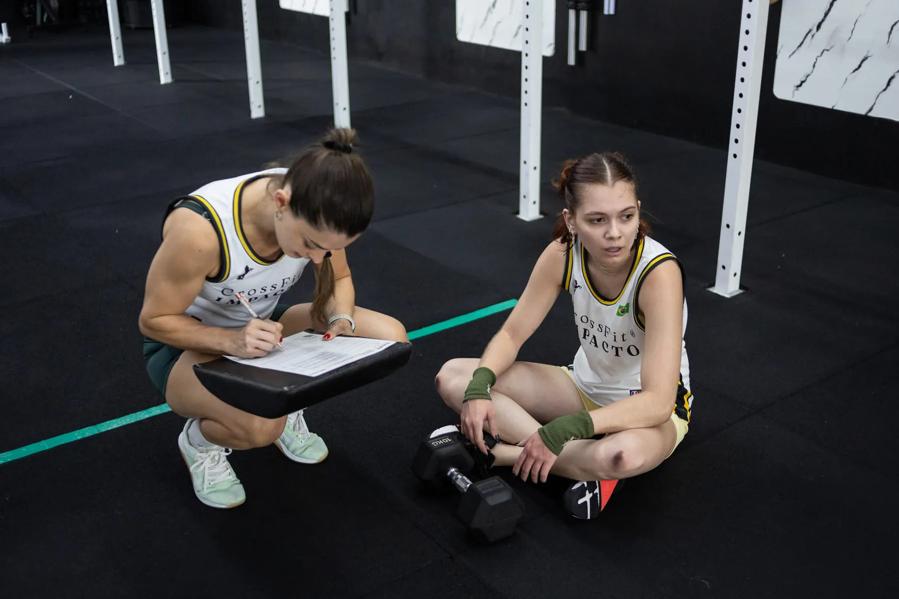Coach agachada anotando na prancheta ao lado de uma atleta sentada no chão, com o halter ao lado.